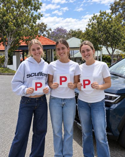 Friends Daisy, Liv and Ashlyn smile and hold up their red P plates in front of a blue car