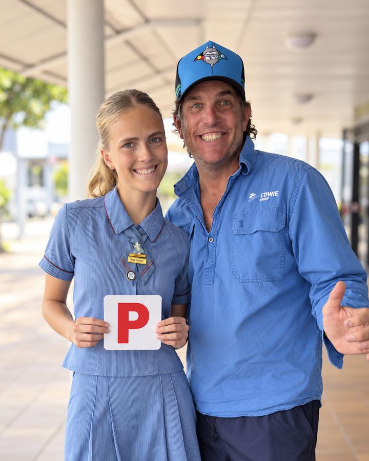 A smiling schoolgirl holds up her red P plate beside her proud dad, who gives a thumbs up, after passing her driving test with A Safe Start